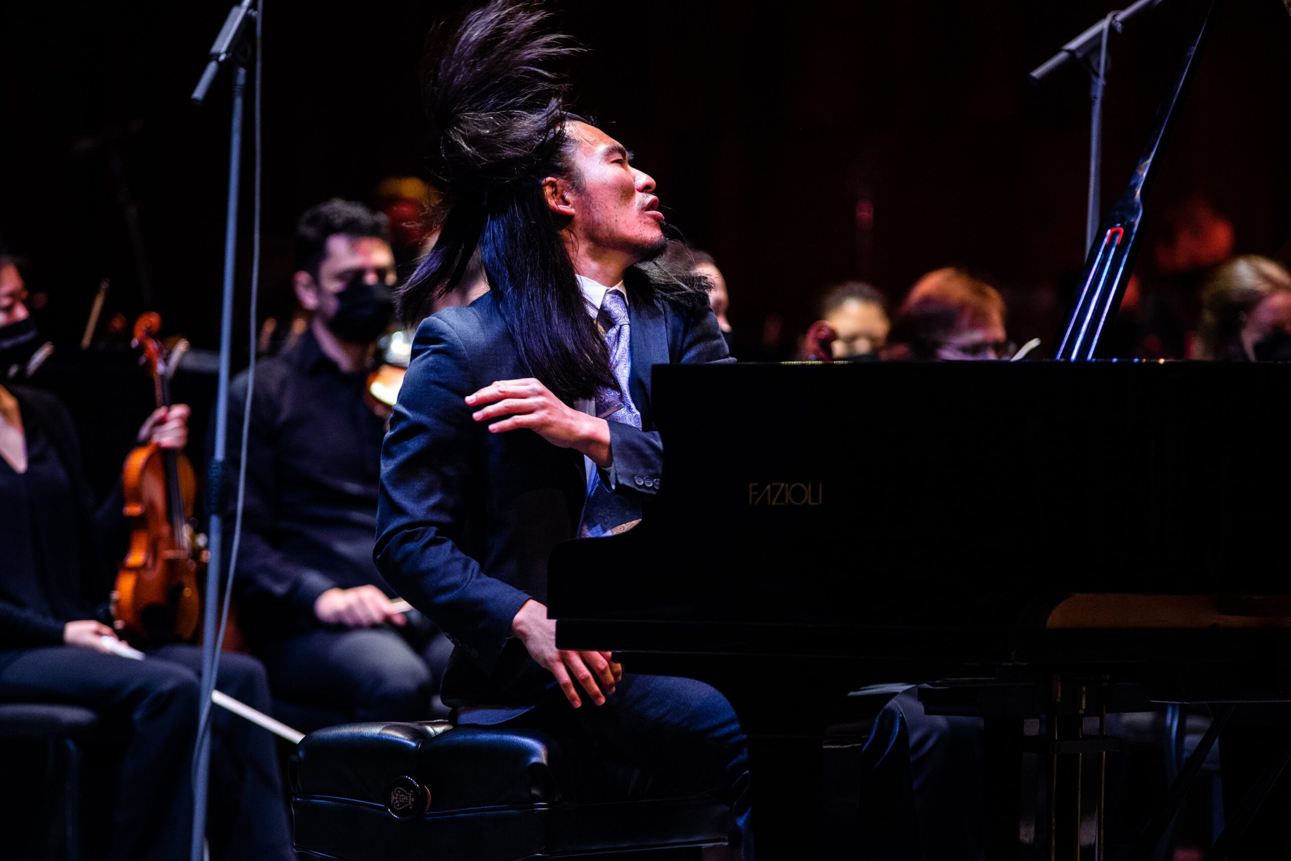 Sebastian Chang throws back his hair in a dramatic gesture while playing a virtuostic passage on a Fazioli Grand Piano onstage in front of the Britt Festival Orchestra premiering his Piano Concerto "The Empress". He is dressed in a dark grey suit and is wearing a purple tie with a tie clip. His hair is styled half up, half down, and is flying in the air.